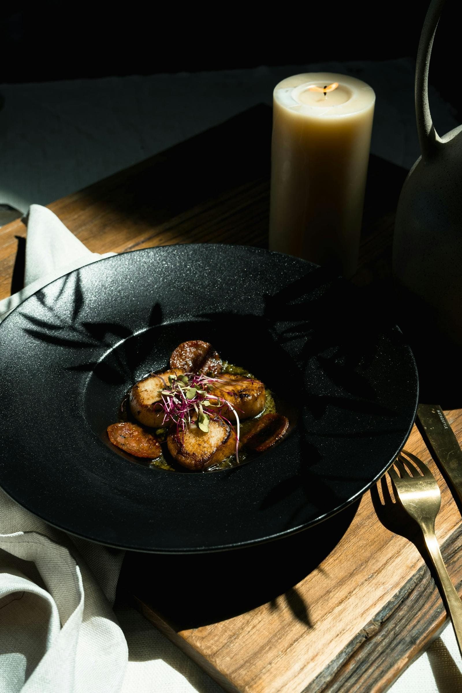 A black ceramic plate of seared dish on a wooden board with a pillar candle in low warm light